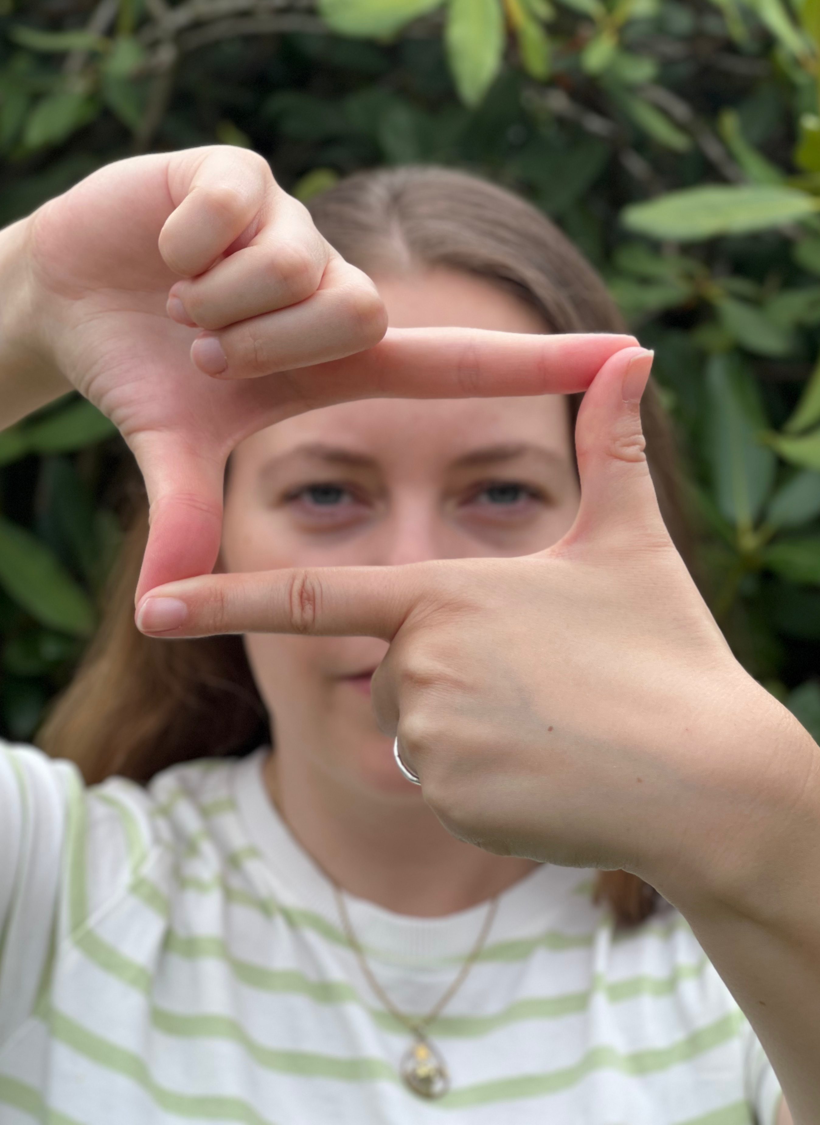 Eine Frau bildet mit ihren Händen einen rechteckigen Rahmen vor ihrem Gesicht, durch den sie hindurch schaut. Die rahmenden Hände sind scharf und die Person unscharf erkennbar.