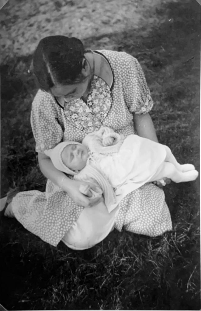 Das Bild ist eine Schwarzweiß-Fotografie. Eine Frau in einem Kleid sitzt im Gras. Sie schaut auf ein schlafendes Baby, das auf ihrem Schoß liegt. Das Baby trägt ein weiße Mütze, Wolljacke, Kleid und Strumpfhose.
