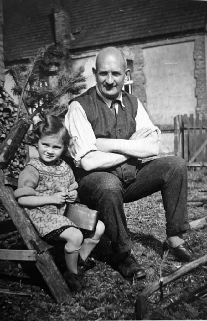 Das Bild ist eine Schwarzweißfotografie. Ein älterer Mann sitzt neben einem kleinen Mädchen im Freien, beide lächeln leicht. Der Mann trägt eine Weste und hat die Arme verschränkt, während das Mädchen in einem Kleid sitzt und ein kleines Behältnis auf dem Schoß hat. Im Hintergrund sind Bäume und ein Gebäude sichtbar.